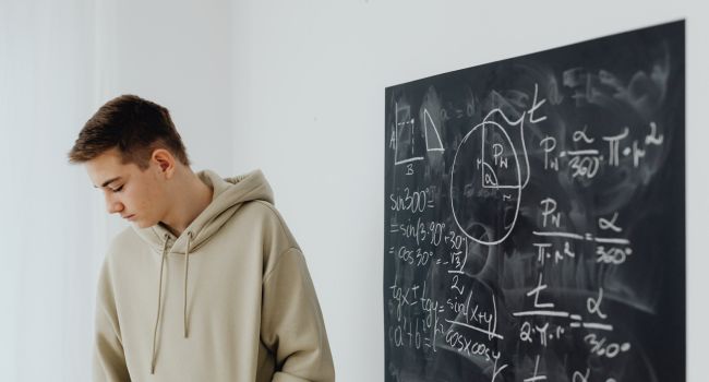 Teen boy looking down beside a chalkboard filled with math equations, representing the challenges and common reasons, such as academic struggles or behavioral issues, that may lead to school suspension.