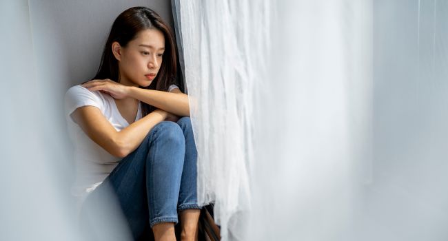 Teen girl sitting alone by a window, appearing withdrawn and thoughtful, representing signs of depression often experienced by teen girls, highlighting the need for awareness and support.