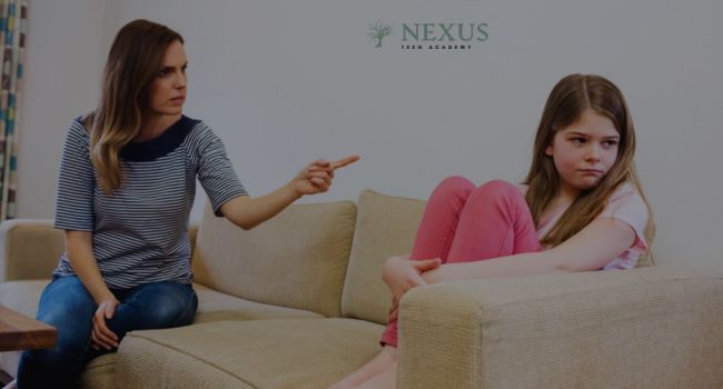 A mother sitting on a couch pointing at her teenage daughter, who looks away upset, representing the dilemma parents face when deciding whether to punish their teen for vaping.