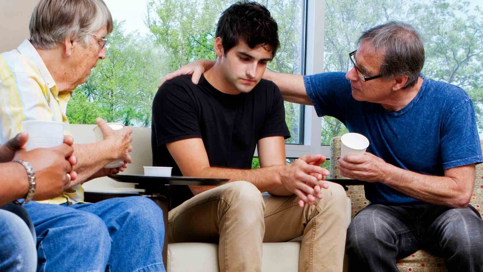 A distressed teen sits in a support group as an older man comforts him, highlighting the importance of early intervention in addressing teen psychosis and mental health.