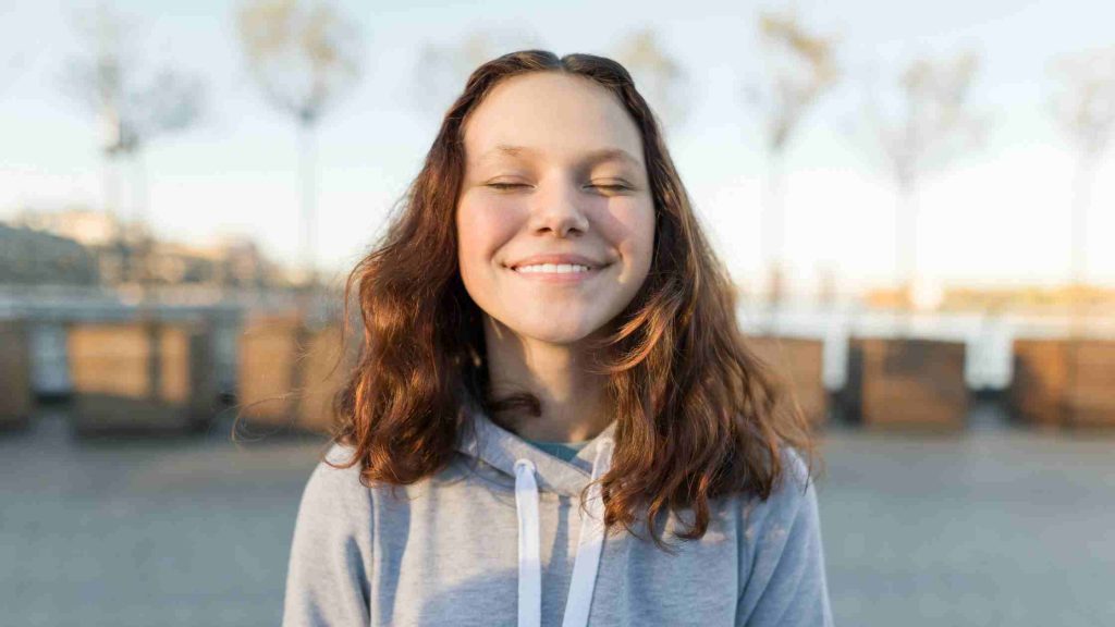 Smiling teen girl with closed eyes enjoying a peaceful moment outdoors, representing the key benefits of mindfulness for teens in reducing stress and enhancing focus.