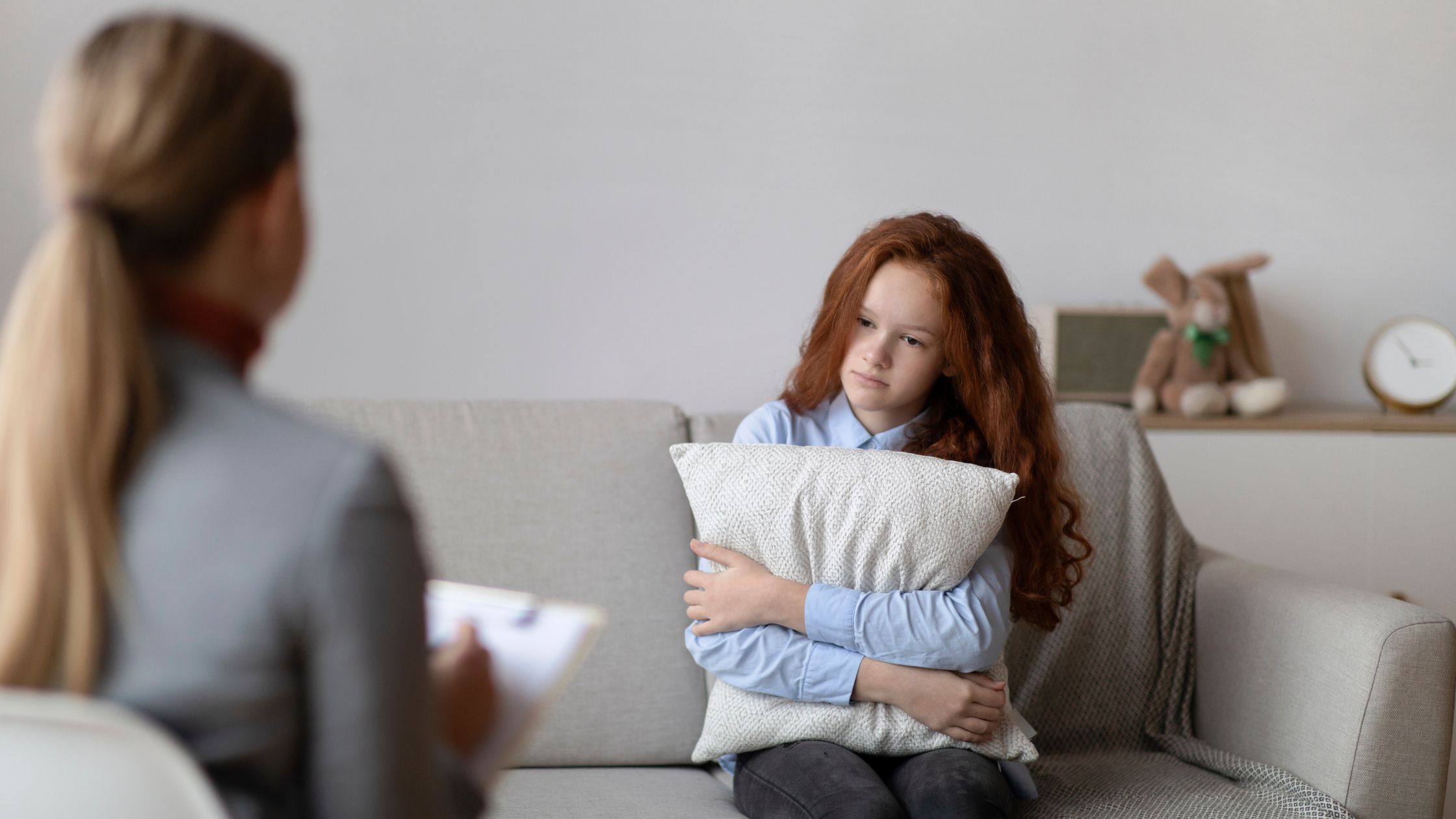 Teen girl with red hair hugging a pillow while talking to a therapist in a cozy setting. Represents behavioral health treatment at Nexus Teen Academy.