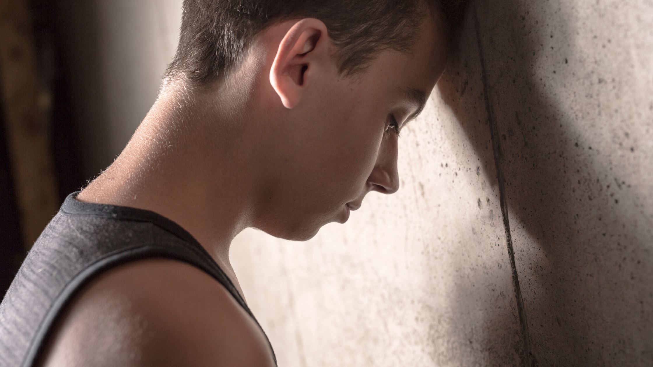 A teenage boy in a dark tank top leaning his head against a concrete wall, looking distressed, symbolizing emotional struggles and risk factors for BPD.