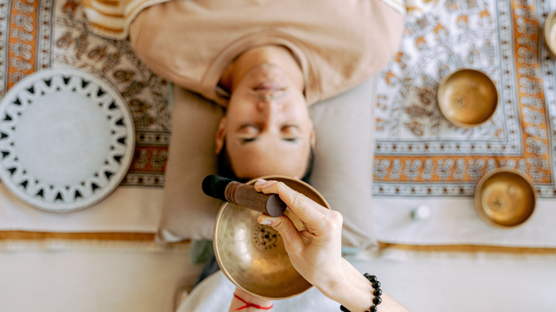 Person lying down with eyes closed during a sound healing session, as a practitioner holds a Tibetan singing bowl above their body, creating soothing vibrations.