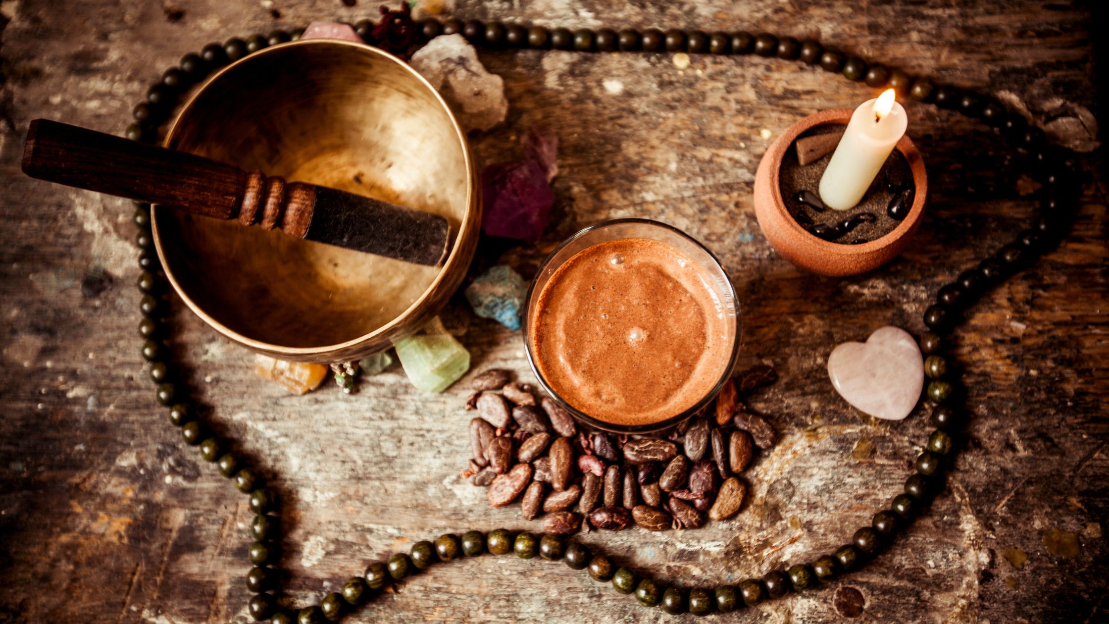 Singing bowl, cacao, crystals, candle, and mala beads arranged on a rustic wooden surface for sound healing and meditation practices.