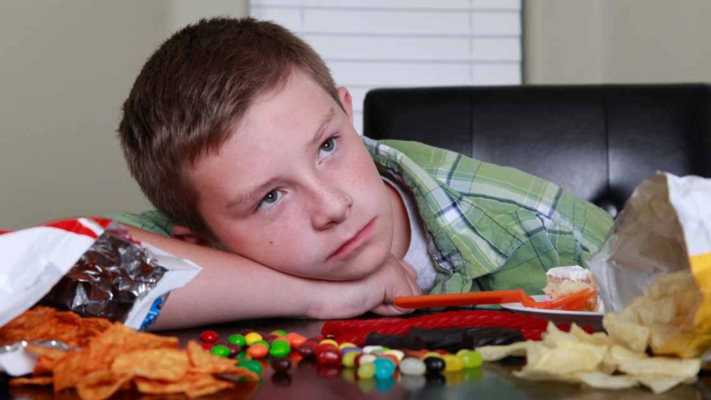 Sad teen boy resting his head on a table surrounded by junk food, symbolizing common causes of eating disorders in teen boys.