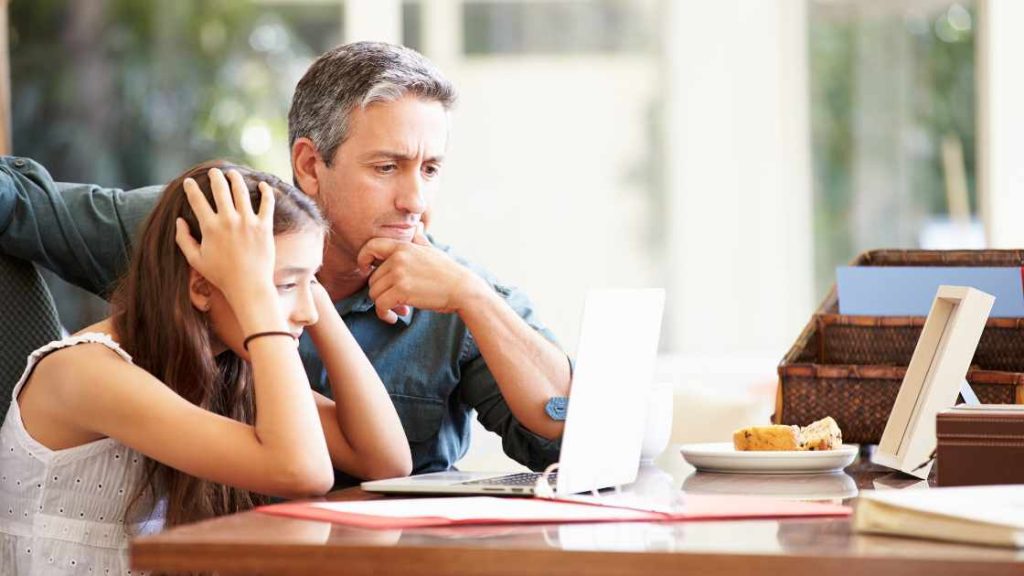 Concerned father supporting stressed teen daughter while looking at a laptop, illustrating factors influencing teen behavioral health.