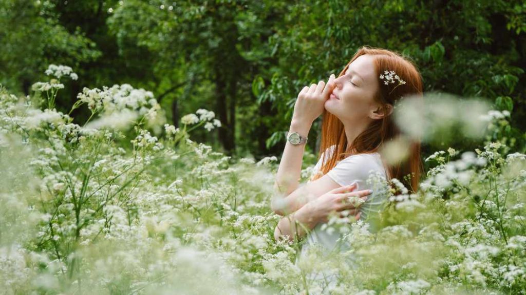 A young woman peacefully embraces herself while standing in a meadow, eyes closed and surrounded by wildflowers, symbolizing the gentle, self-compassionate practice of Loving-Kindness Meditation (Metta).