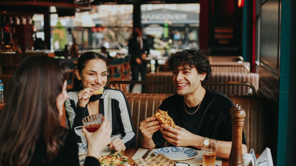Group of friends laughing and eating at a trendy restaurant, capturing social dynamics and cultural influences around food and body image.
