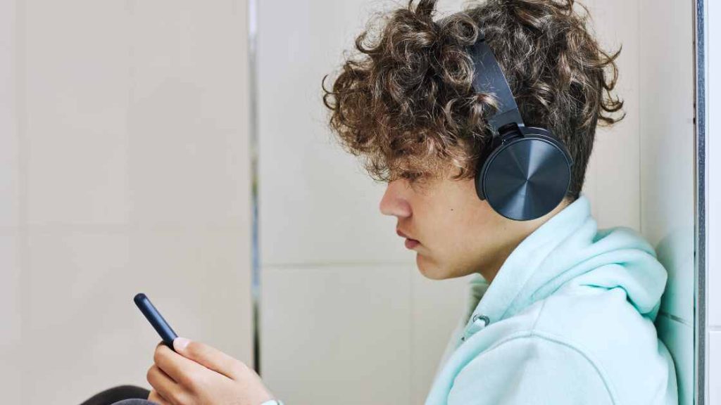 Teen boy in headphones looking at phone with serious expression, illustrating the impact of eating disorders on teen boys.