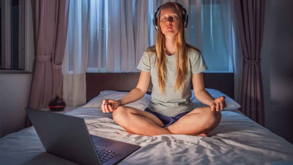 Teen girl practices meditation on her bed at night, wearing headphones and using a laptop, highlighting accessible and calming top meditation exercises for teens in a digital, cozy setting.
