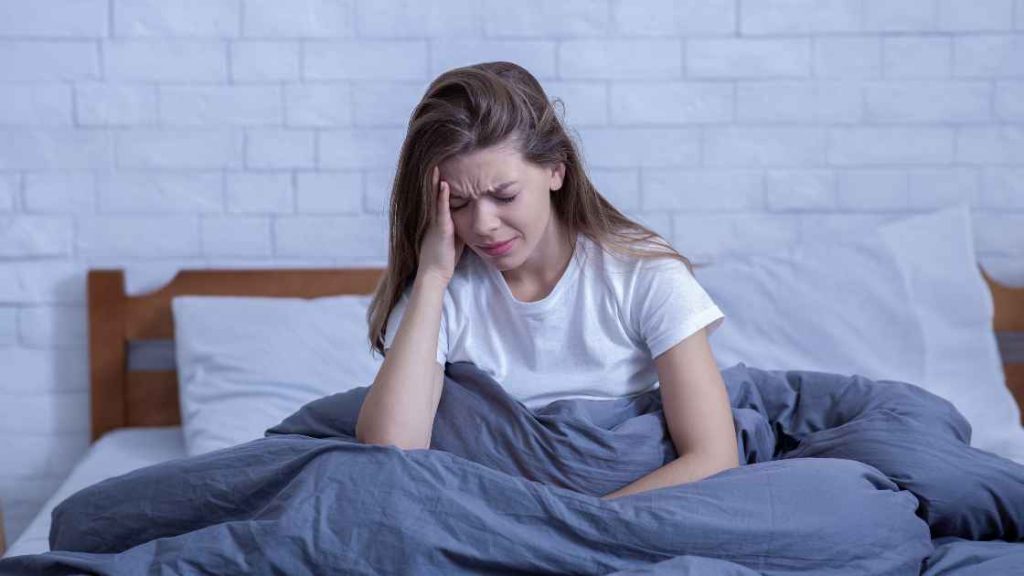 A distressed teenage girl sits in bed with her hand on her head, representing emotional struggle and the need for inpatient treatment to address PTSD symptoms in adolescents.