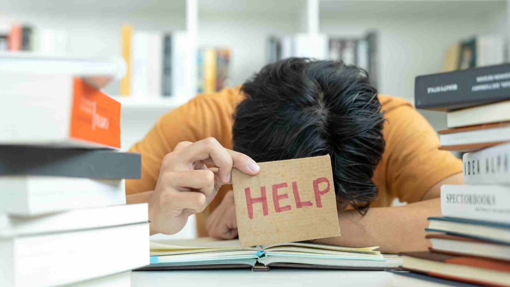 Teen overwhelmed by social phobia hides face on desk, holding a "HELP" sign among stacks of books.