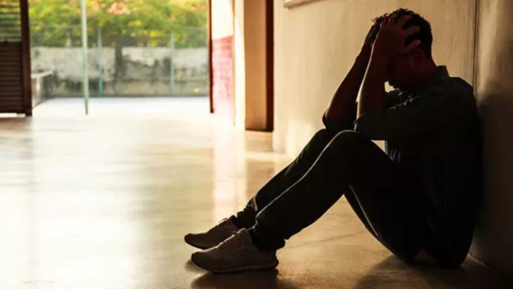Teen boy sitting alone against a hallway wall, experiencing grief and rising rage after a painful breakup.